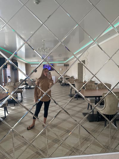The image shows a reflection in a diamond-patterned mirror wall of a well-lit, modern interior. The reflection reveals a woman taking a selfie with her phone. She is wearing a light brown hooded sweatshirt and black leggings, with black flip-flops. Her hair is long and blonde. The background of the reflection shows a dining area with a long wooden table and several tufted beige upholstered chairs. In the distance, there is a chandelier and some accent lighting on the ceiling. The flooring appears to be a tiled surface with a wood-grain pattern. Another reflection shows a smaller child looking at the camera from further back in the room. The overall impression is one of a contemporary indoor space, possibly a restaurant or a spacious home, during daylight hours. The mirrored surface distorts the reflections, creating a mosaic-like effect.