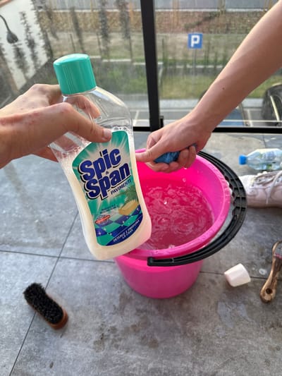 Two individuals are in the process of cleaning. One person is holding a bottle of Spic & Span floor cleaner over a bright pink bucket filled with soapy water. The other person is wringing out a blue sponge into the same bucket. The scene is set on a tiled floor, likely outdoors or in a sunroom, with a blurred background suggesting a parking lot and some vegetation. Scattered items include a small brush, a bar of soap, and a plastic water bottle. The lighting indicates it is daytime. The image does not contain any visual information that would allow for the determination of the location within Białystok, Poland.
