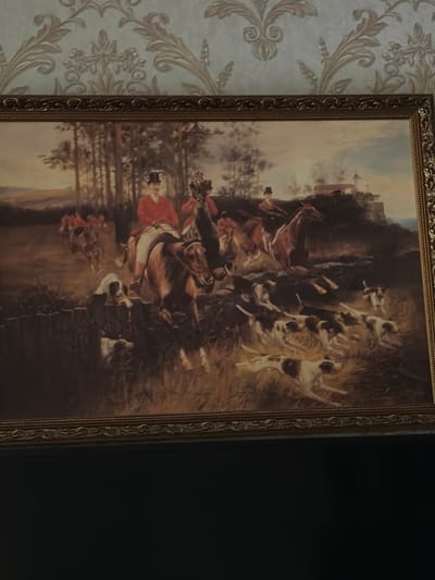 The image is a painting depicting a fox hunt. In the foreground, several dogs are running through tall grass. Behind them, two men on horseback, dressed in red coats and top hats, are also riding. One horse is mid-leap over a fence. In the background, more riders and horses are visible, as well as a wooded area and a building on a hill. The painting is framed in an ornate gold frame. The painting is hanging on a wall with patterned wallpaper. There is no indication of Tambov, Russia in the image.