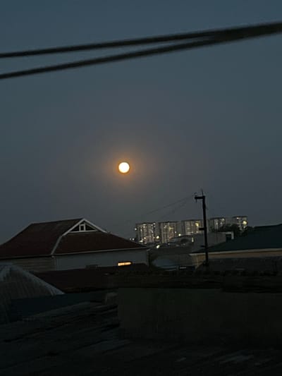 The image shows a dusky sky over what appears to be a residential area. A bright, round moon is prominently visible in the upper center of the frame. Below the moon, a house with a gabled, reddish-brown roof and a small triangular window is in the foreground. In the mid-ground, there are other buildings and structures, including a utility pole with wires. In the background, a cluster of tall buildings with illuminated windows is visible, suggesting an urban or suburban setting. The overall atmosphere is dim, consistent with either twilight or a hazy night. The image is taken from a slightly elevated perspective, looking out over rooftops. There are no people visible in the image. The location is identified as Sabunchu, Azerbaijan.