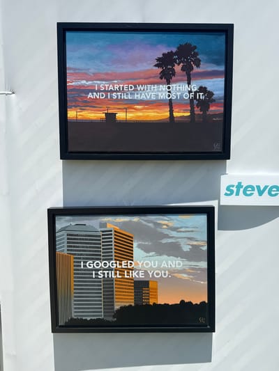 The image shows two framed art pieces hanging on a white wall, possibly in an art gallery or a home. Above the frames, there is a vertical white sign with the name "steve" printed on it in light blue.
The art piece on the left depicts palm trees silhouetted against a sunset sky. White text is superimposed on the image, reading, "I STARTED WITH NOTHING AND I STILL HAVE MOST OF IT." The frame is black.
The art piece on the right shows buildings silhouetted against a sunset sky. White text is superimposed on the image, reading, "I GOOGLED YOU AND I STILL LIKE YOU." This frame is also black.
The lighting in the image indicates a strong light source, causing the surfaces to cast shadows, and the photo appears to be taken indoors. The location is Beverly Hills, United States of America.
