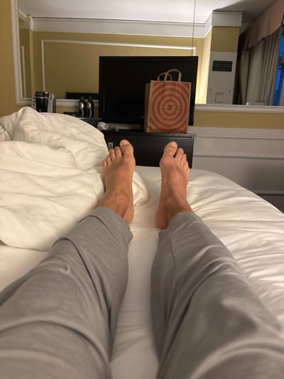 The image shows a person's legs and feet in grey pajamas, resting on a white bed. The perspective is from the person's point of view, looking down their legs. In the background, a black television is visible on a dark surface, with a red and white striped shopping bag on top of it. A coffee maker and two bottles are also present on a shelf to the left of the television. The room appears to be a hotel room, with yellow walls and white molding. The image was taken indoors. No specific location in San Francisco, United States of America is indicated by the image content.