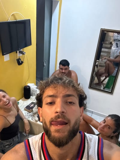 A selfie captures four individuals in a casual setting. One woman smiles in the background. A shirtless man appears behind the selfie taker. Another woman is positioned in the lower part of the frame. A television sits on a yellow wall next to a closed grey door. A framed image hangs high on the wall featuring two people. A black speaker, book, and assorted items rest on a table.