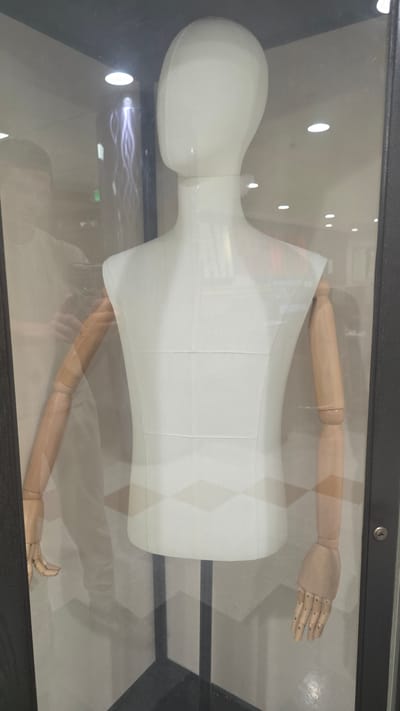 The image shows a headless white fabric mannequin with articulated wooden arms and hands, displayed behind glass. The mannequin is positioned centrally in the frame, with its torso a plain white fabric shape and its arms extending outwards. The left arm is partially obscured by a reflection of a person holding a phone, who appears to be looking at the mannequin. The right arm is fully visible, ending in a wooden hand with articulated fingers. The background visible through the glass includes a wall with a geometric pattern, lighting fixtures, and a dark vertical frame on the right side of the image, suggesting the display is inside a shop or exhibition space. The overall scene is indoors and well-lit.