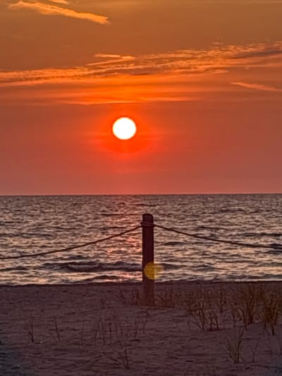 This is a photograph of a sunset over a beach in Wasaga Beach, Canada. The sun is a bright orange orb low on the horizon, casting a warm glow across the sky and water. The sky is a gradient of orange and red, with some wispy clouds. The ocean is calm, with gentle waves reflecting the colors of the sunset. In the foreground, there is a sandy beach with some dry grass and a wooden post with a rope strung between it and another unseen post, suggesting a boundary or railing. The overall impression is one of tranquility and natural beauty.