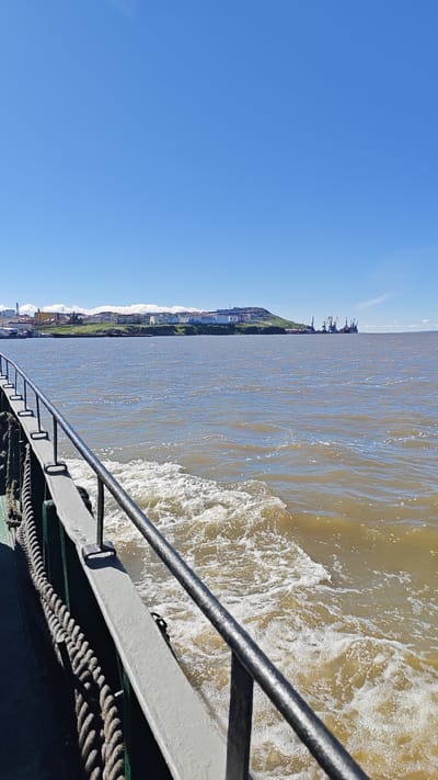 A vertical, medium shot captures a view from a vessel moving through choppy, brown water under a clear, bright blue sky. In the foreground, the dark green hull of the vessel is visible on the left, with a thick, coiled rope secured to a railing. The railing itself is a dull gray metal, featuring horizontal bars and vertical supports. White foam churns in the wake of the vessel, creating a turbulent pattern in the water.

In the mid-ground and background, a coastal city is visible on a green, hilly landscape. The city features a collection of buildings with a variety of colors and architectural styles, suggesting a developed urban area. Some of these buildings are white, while others display hues of yellow, orange, and blue. On the right side of the mid-ground, industrial structures, including tall cranes, are present, indicating a port or industrial zone. The overall impression is of a sunny day with moderate sea conditions. The scene depicts a view of Anadyr, Russia, from the water.
