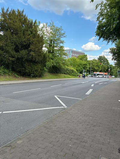 The image shows a street in Aachen, Germany. On the right side of the road is a paved sidewalk. The road has white lane markings and a center line. On the left side of the road is a grassy area with trees and bushes. In the background is a building with red and orange accents. Further up the road, two pedestrians wearing reflective vests are walking. Behind them is a bus at a red light. Traffic lights are visible. The sky is blue with some clouds, and trees are visible overhead.