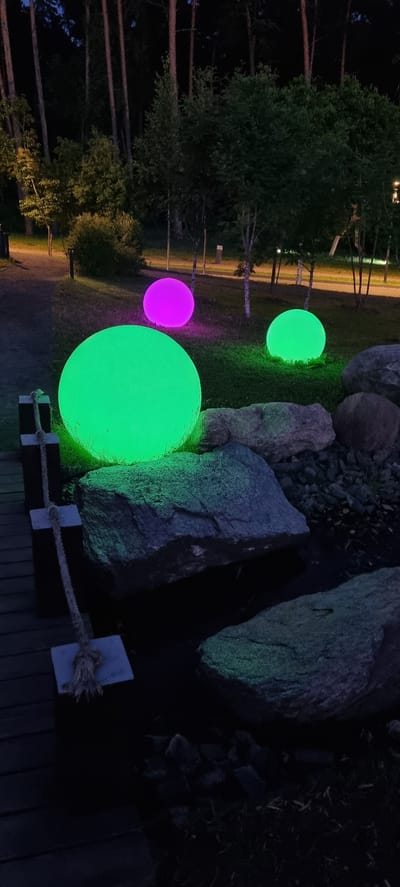 The image depicts an outdoor scene at night, illuminated by several large, glowing orbs. These orbs are colored pink and green, casting a vibrant glow on the surrounding grassy area and rocks. The largest orb, a bright green, is positioned in the foreground. Behind it, a smaller pink orb is visible, and further back, another smaller green orb.

The setting appears to be a park or garden, with mature trees visible on the left side of the frame, their trunks silhouetted against the dark background. The trees suggest a wooded or forested area. In the background, a paved pathway or road is illuminated by subtle lighting, with thin tree trunks lining it. Large, rough-textured rocks are also a prominent feature in the scene, especially on the right side, some of them catching the colorful light from the orbs.

A rustic wooden bench is partially visible in the lower right corner, with a rope barrier made of braided material and small wooden posts. The rope is secured to the bench and also to a square wooden post. The overall ambiance is serene and somewhat magical, with the glowing orbs creating an enchanting atmosphere. The time of day is clearly night, and no specific weather conditions are evident, but the darkness suggests clear skies. The location cues, such as the type of trees and the general landscape, are consistent with a natural outdoor environment. There is no visible text in the image.