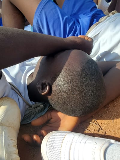 The image shows a close-up, overhead view of a person lying down, with their head tilted to the side. They are wearing a white t-shirt and blue shorts. Their arms are bent, and their hands are resting on their head, possibly to shield their eyes from the sun. The person's skin tone is dark, and their hair is short and dark. White sneakers are visible in the foreground and to the right of the frame. The ground beneath appears to be sandy or dusty, with some small sticks scattered on it. The lighting suggests it is daytime, with strong shadows cast by the person's head. The scene seems to be outdoors, possibly in a rural or open area given the ground texture. There is no discernible text in the image.