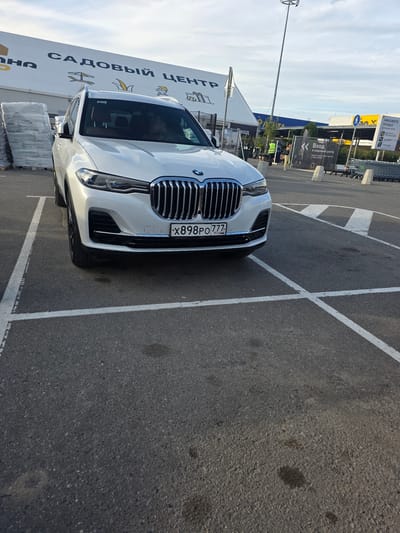 This media file shows a white BMW parked in an outdoor parking lot in Sholokhovo, Russia. The car is positioned at an angle, facing towards the left of the frame, with its front grille and license plate clearly visible. The license plate reads "X898PO 777 RUS".
In the background, a large building with signage is visible. The signage on the building includes Russian text that appears to indicate a "Garden Center" ("САДОВЫЙ ЦЕНТР"). Other signs nearby mention "Entrance to the garden center" ("Вход в садовый центр") and "Order a project for your home" ("ЗАКАЖИТЕ ПРОЕКТ для вашего дома").
The parking lot is paved with asphalt and marked with white lines, indicating parking spaces. Several shopping carts are visible lined up in the distance. The weather appears to be clear and sunny, suggesting it is daytime, likely in the late afternoon or early evening given the angle of the light. The overall scene depicts a commercial area, likely a large retail or garden center, with a vehicle parked in its designated area. There are no people prominently featured in the image, and the focus is on the car and the surrounding environment.