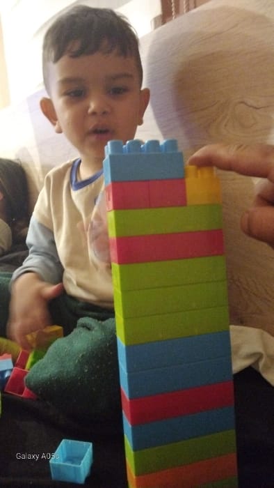 The scene is depicted as occurring indoors in Oreshene, Bulgaria. A young child, with dark hair and light skin, is positioned in the left half of the frame, looking forward with a partially open mouth. The child wears a light-colored long-sleeve top with blue trim at the collar and dark legwear, with a green sock visible on the left foot. In the central foreground, a vertical tower approximately 14 blocks high, constructed from interlocking plastic building blocks, stands prominently. The blocks are arranged in alternating colors including green, blue, red, orange, and yellow. An adult's hand, entering the frame from the upper right, is captured in the act of placing a single yellow block onto the uppermost section of the tower. Scattered on a dark surface in the lower-left foreground are additional loose plastic blocks, including red, blue, and yellow elements, alongside a distinct light blue hollow square block. The background is blurred, featuring a light-toned surface with a wood grain pattern on the right and an indistinct, soft, light-colored upholstered surface on the left.