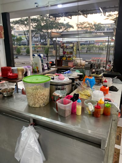 The image depicts a food stall counter, likely located in Mekar Bakti, Indonesia, given the context. The counter is made of stainless steel and is cluttered with various food-related items. In the foreground, a large transparent plastic container filled with what appear to be small fried snacks is prominent. To its left, a metal bowl contains a dark sauce. Several smaller containers holding sauces or condiments, as well as small plastic baskets with black utensils, are arranged on the counter. In the background, a large cooking pot with a pink lid is visible, along with blenders and other kitchen equipment. The stall appears to be set up outdoors, with trees and buildings visible through an open-air structure. The lighting suggests it might be late afternoon or early evening. There are no people visible in the frame.