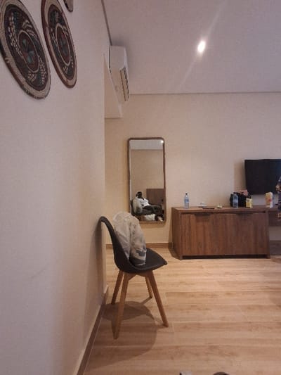 The image displays the interior of a modern room. The walls are light-colored, and the floor is covered with light wood-patterned tiles. On the upper left wall, two circular, woven decorative plates with intricate patterns are mounted. A white wall-mounted air conditioning unit is affixed high on the left wall, near the ceiling.
In the mid-ground, a dark gray chair with light wooden legs rests against the left wall, supporting a translucent white plastic bag on its seat. To the right of the chair, a rectangular mirror is mounted on the wall, reflecting a person seated on an unseen surface, possibly a bed or sofa. The individual in the reflection appears to be wearing light-colored clothing.
Further to the right, a low, rectangular wooden cabinet with two doors is positioned against the wall. On its surface, a blue water bottle, a dark bottle, and other containers are arranged. A black flat-screen television is mounted on the wall above the cabinet. A small portion of patterned fabric or a curtain is visible at the extreme right edge of the frame. The ceiling is light-colored and features a single recessed light fixture. No discernible elements within the image identify the specific location as Takum, Nigeria.