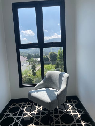 The image displays a sunlit corner of an interior room located in Kayuambon, Indonesia. A large, black-framed window, divided into four main sections, dominates the scene, offering an expansive view. A light grey upholstered armchair with slender dark legs is positioned in the corner, angled towards the window. The room features plain white walls meeting at a corner and a black carpet with a repeating white geometric pattern of intersecting lines. A black baseboard borders the floor. Strong sunlight streams through the window, casting sharp shadows across the armchair and the patterned carpet. The upper sections of the window reveal a blue sky with scattered white cumulus clouds and distant green hills. The lower sections present an outdoor landscape including a white building with a reddish-brown roof, surrounded by various green trees and vegetation, and what appears to be terraced hills in the background. A paved surface is visible in the immediate outdoor foreground.