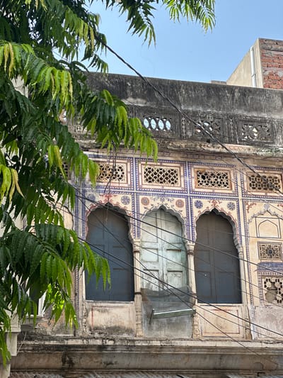 This image depicts the exterior of a traditional building in Pushkar, India, characterized by its ornate architecture. The facade features arched windows with dark wooden shutters, framed by intricate decorative patterns in blue and red. Above the windows, there are several smaller square openings with latticework, possibly serving as ventilation or decorative elements. The building appears aged, with weathered plaster and some signs of decay, but retains its historical charm. Lush green foliage from a tree in the foreground partially obscures the view of the building, adding a natural element to the urban scene. Overhead, electrical wires crisscross the frame, indicative of the modern infrastructure overlaid on the historic setting. The sky above is a clear blue. The scene captures a blend of old-world architecture and everyday life in an Indian city.