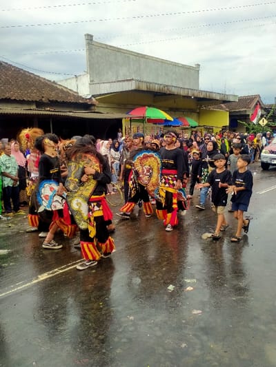 The image depicts a cultural procession moving along a wet asphalt road in the city of Mojogedang, Indonesia, under an overcast sky. Approximately six individuals in the foreground and midground are dressed in elaborate traditional Javanese attire, consisting of black tops, red sashes, and yellow-orange patterned leg wraps. Each carries or is integrated with a stylized, colorful horse effigy or shield. One performer on the far left wears a large, yellow, lion-like headpiece. All performers are walking from left to right. Two young boys in dark t-shirts and shorts walk parallel to the procession on the right. A crowd of spectators, including many individuals wearing headscarves, lines both sides of the street, observing the event. In the background, a single-story building with a dark tiled roof is visible on the left, and a larger, light-colored multi-story building with a flat roof is on the right. Several multi-colored market umbrellas are visible behind the spectators on the right, along with an Indonesian flag mounted on a pole. A light-colored sedan is partially visible on the far right. The wet road surface reflects light and shows faint white markings and scattered small debris.