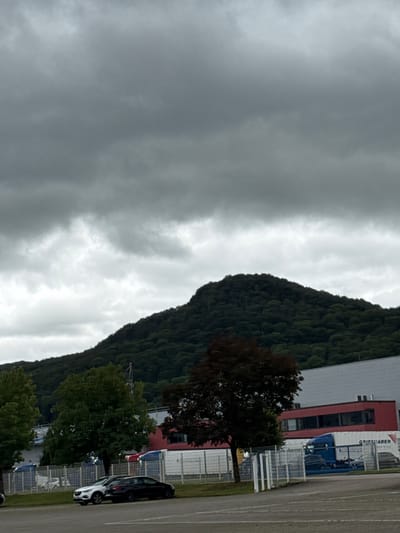 The image depicts an overcast day with heavy, dark clouds dominating the sky. In the foreground, a paved area, likely a parking lot, is visible. Two cars are parked side-by-side on the grass verge next to the parking lot: a white compact SUV and a dark-colored sedan. Behind the cars, a fence separates the area from what appears to be an industrial or commercial zone.

Beyond the fence, a long, multi-story building with a red upper section and a lighter gray lower section is visible. Several large trucks, including a blue semi-trailer truck with "GRIESHABER" written on its side, are parked near the building. Green trees are scattered throughout the scene, with a dark-leafed tree prominently positioned in the middle ground.

In the background, a densely forested hill or small mountain rises, its slopes covered in dark green foliage. The overall impression is one of a somewhat industrial landscape situated at the base of a natural, wooded area. The location is identified as Bad Säckingen, Germany.