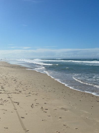 The image depicts a wide, sandy beach with gentle waves rolling in from the ocean under a clear blue sky with scattered clouds. The foreground shows a textured sand surface with numerous footprints and a distinct tire track pattern from a vehicle. In the middle ground, the ocean is a deep blue-green, with white foam from the breaking waves. A few distant figures, appearing as small silhouettes, are visible along the shoreline and in the water. The horizon line separates the ocean from the sky, with a thin line of clouds in the distance. The overall impression is a serene, natural beach scene.

While the image captures a typical beach environment, the specific location within Surfers Paradise, Australia, cannot be determined from the visual information alone. Surfers Paradise is known for its wide, golden beaches and the consistent surf, which aligns with the scene depicted. However, without any identifying landmarks or signage, pinpointing the exact spot within Surfers Paradise is not possible based on the provided image.