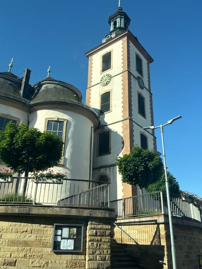 The image depicts the St. Andreas Catholic Church in Ubstadt, Germany. The photograph is taken from a low-angle perspective, showcasing the church's tower and dome against a clear blue sky. The tower is rectangular with a clock on one of its faces and a pointed spire. The dome is topped with decorative elements. The church's exterior is light in color with contrasting darker trim. A street light is visible near the tower. The church is adjacent to a stone wall with a metal railing and stairs. A framed bulletin board containing notices and flyers is mounted on the wall. Green bushes are growing around the church's lower levels.