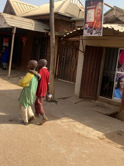 The image captures a street scene in Kaduna South, Nigeria, under bright daylight. In the foreground, two young, bald-headed males, likely children, walk from right to left. The male on the right wears a red short-sleeved top and maroon trousers with light-colored flip-flops; he carries a small, spherical, metallic bowl in his left hand and has his right arm around the shoulder of the male on the left. The male on the left wears a yellow and light-green short-sleeved top, light-colored long trousers, and dark-colored sandals.
To the right of the subjects, a building features a brown wooden door and a glass storefront displaying multiple large posters of women, including a prominent vertical banner reading "DOBMAKEOVER" with an image of a woman in a headwrap and details about beauty services. A tall, unpainted wooden utility pole stands centrally, between the walking subjects and the right-side building. Behind this pole, a large wooden gate with dark metallic decorative elements is visible, and barbed wire is affixed to a structure above it. In the far-left background, another building with a corrugated metal roof and an awning supported by wooden poles is partially visible, with a person in a blue garment standing near its entrance. The ground surface consists of light-brown dirt and uneven patches of concrete or asphalt. Strong sunlight casts distinct shadows across the scene, indicating a bright, clear day.