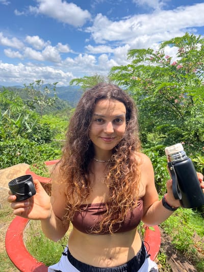 A woman stands outdoors with a lush green landscape and cloudy blue sky behind her. She is holding a black thermos in her right hand and a black lid in her left hand. She is wearing a brown strapless top and white and black shorts. The location appears to be a scenic overlook in Son Dien, Vietnam, with rolling hills and vegetation visible in the background.