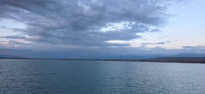 This is a wide-angle, landscape photograph taken from a body of water, likely Lake Issyk-Kul, near Pristan'-Przheval'sk, Kyrgyzstan. The image captures a serene, overcast scene at dusk or dawn.
The foreground is dominated by the tranquil, blue-green water of the lake, which exhibits a gentle ripple pattern across its surface. The horizon line is clearly defined, separating the water from the land and sky.
In the mid-ground, a strip of land is visible, lined with trees and low-lying buildings that suggest a settlement or town. The land appears somewhat barren or dry. Beyond this, a range of mountains stretches across the width of the image, their forms softened by distance and atmospheric haze. These mountains are layered, with the furthest peaks appearing a hazy blue against the sky.
The sky is filled with heavy, dark gray and blue clouds, typical of an overcast day. Patches of lighter sky, possibly indicating the direction of sunset or sunrise, are visible through breaks in the clouds. The overall color palette is muted, dominated by blues, grays, and subdued earthy tones. There are no people or animals visible in the scene. The image conveys a sense of peaceful solitude and the vastness of the natural landscape.