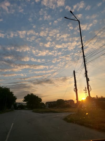 The image depicts an exterior scene at dusk in Strazhitsa, Bulgaria, characterized by a low angle view of the sky, utility poles, and a road. The sky is a prominent feature, filled with a pattern of fluffy, light-colored clouds against a blue background, with the sun's golden rays breaking through the horizon. Two tall utility poles with multiple wires are silhouetted against the sky, one with a double lamp fixture at its apex. The foreground shows a paved road that curves to the right, with a white line marking its edge on the left. To the right of the road, overgrown grass and vegetation are visible, leading towards buildings in the middle ground and background. These buildings appear to be residential, with a few multi-story structures visible. The overall impression is one of a quiet, possibly rural or suburban, street in Strazhitsa during sunset. There are no visible people or animals in the frame.