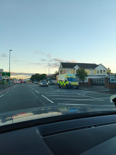 The image shows a street scene in the city of Sandwell, United Kingdom, likely during the late afternoon or early evening. The sky is a pale blue with some wispy clouds and a sliver of a crescent moon visible. In the foreground, the dashboard and windshield of a vehicle are visible, reflecting the street and surrounding environment.
On the road, there is traffic, with several cars and an ambulance. The ambulance, a yellow and green vehicle with flashing blue lights, is positioned on the right side of the road, appearing to be stopped or moving slowly. Several cars are also on the road, some in traffic lanes and others in the process of turning.
To the left of the road, there is a blue lamppost and a signpost for "Esso" and "Morris." Further down the road, traffic lights are visible, some of which are red, indicating traffic is stopped.
On the right side of the road, there is a yellow building with a dark roof, possibly a commercial establishment or residential building. A black metal fence is also visible in front of this building. Trees are present in the background, and the overall environment suggests an urban or suburban setting. The scene appears calm, with no immediate indication of an emergency situation beyond the presence of the ambulance.
