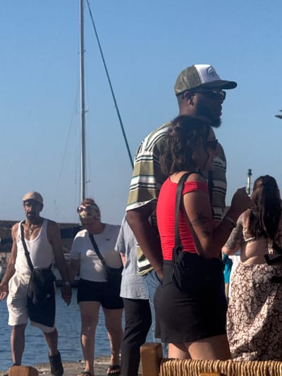 The image shows a group of people standing outdoors near a body of water, likely a harbor, given the presence of a sailboat mast in the background. The scene appears to be in a city, with buildings faintly visible in the distance. The sky is clear and blue, suggesting a sunny day.
In the foreground, a man wearing a white tank top, white shorts, and a tan flat cap stands to the left, with a black messenger bag slung across his chest. He is looking to his left. Next to him, a woman in a white t-shirt and black shorts, with a bandana on her head, is looking down. To the right of these two, a man in a striped shirt and a baseball cap is partially visible. In front of the man in the striped shirt, a woman wearing a red off-the-shoulder top and a black skirt is turned to her right, with tattoos visible on her arm. She has a black crossbody bag. Further to the right, another woman in a floral-patterned dress is visible, facing away from the camera.
The people are standing on a paved surface, possibly a boardwalk or dock. The water is visible in the lower left portion of the image. The overall impression is a casual outdoor gathering in a coastal urban setting. This scene is consistent with the waterfront areas of Chania, Greece, known for its Venetian harbor and bustling promenade.