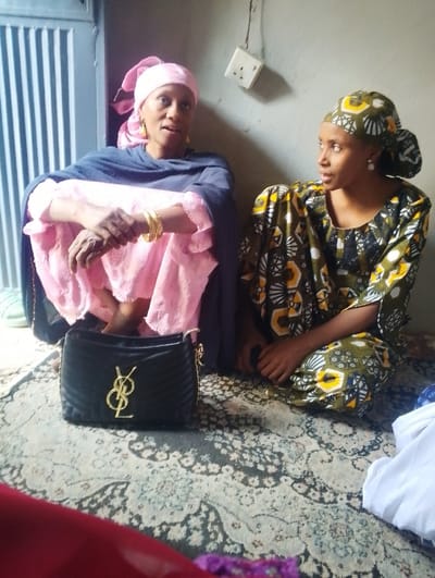 Two adult females are seated on a patterned rug in an indoor setting in Dorayi, Nigeria. The woman on the left, appearing older, wears a pink headscarf, a pink garment, and a dark blue shawl. Her bare feet are partially visible, and she has multiple gold-colored bangles on her right wrist and a gold earring on her visible left ear. Her mouth is slightly open, indicating she is speaking. The woman on the right, appearing younger, wears a patterned olive-green, yellow, black, and white headscarf and matching dress, with a pearl-like earring on her visible left ear. She is looking directly at the woman on the left, with her hands resting in her lap. Between them, on the rug, rests a black quilted handbag featuring a gold 'YSL' logo and a chain strap. The background shows a plain, light-colored wall with a white electrical outlet and exposed wiring mounted on it. To the far left, a vertical slatted structure is partially visible. A pair of light green sandals is located near the feet of the woman on the left. White fabric is present on the rug to the far right, and colored fabric is in the immediate foreground.