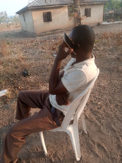 A man in a light-colored shirt and brown pants sits in a white plastic chair, holding a phone to his ear. The background shows a dusty, grassy area with a small, simple house. The house has a corrugated metal roof, and barred windows. The overall setting is rural and somewhat sparse.