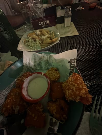 This is a close-up, eye-level shot of a table at a restaurant, possibly located in Pravets, Bulgaria. The table is dark and has a meshed surface. In the foreground, a blue plate holds a serving of chicken wings with a creamy dipping sauce. The wings appear to be breaded and fried. Some of the wings are red, suggesting they are coated in a sauce. A white paper with text is partially visible beneath the wings, some of which is illegible. To the right of the plate, a green plastic fork rests on a white napkin. In the background, a white plate holds french fries and a side salad. To the left, a green bottle of Heineken beer is visible. A dark red box with the Costa Coffee logo is also present. Several empty glasses and a salt shaker are on the table. A person's arm and shoulder are visible in the upper left corner, indicating their presence at the table. The lighting suggests it is evening.