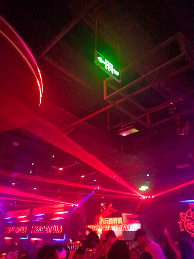 The image depicts the interior of a dimly lit nightclub in Nanning, China, characterized by vibrant red and purple laser lights illuminating the space. A prominent green neon sign with Chinese characters and an arrow pointing to the left is visible, indicating an exit or direction. In the background, a sign reads "YUAN BEAST TAVERN" in English, along with Chinese characters, suggesting the establishment's name. Several patrons are present, with some standing and others blurred in the foreground. The environment appears to be set up for entertainment, with a stage or performance area hinted at by lighting equipment on the ceiling. The overall atmosphere is that of a lively nightlife venue.