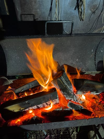 This media displays a vibrant wood fire burning within a dark metal structure, likely an outdoor fire pit or grill. Large wood logs, in various stages of combustion, glow with intense red and orange embers, surrounded by bright, dancing flames in shades of yellow and orange. One prominent flame extends horizontally across the left side of the frame.

The metal structure housing the fire is dark and appears robust. To the left, a bent metal handle or bar is attached to this structure, and further left, a bundle of tangled wires or cables rests on a dark, textured surface, hinting at adjacent equipment. In the background on the right, a dark metal grate or rack is partially visible, subtly lit by the fire's glow.

The overall darkness of the surroundings suggests the image was captured at night or late evening, with the fire being the dominant light source. No individuals or visible text are present in the frame. The scene evokes a sense of warmth and the primal element of fire, set in an outdoor context in Kauchuk Stantsiya, Russia.