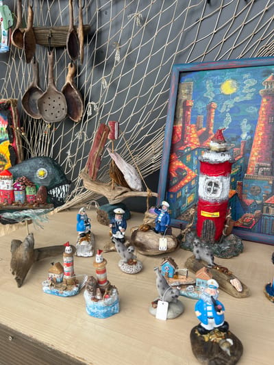 The image displays a collection of nautical-themed decorative items arranged on a wooden surface against a dark gray background, with a fishing net draped behind. Prominently featured is a painting of a cityscape with lighthouses under a moonlit sky, framed in blue. In the foreground, several miniature lighthouses, sailors, and seal figurines are positioned on driftwood and stone bases. One of the lighthouses has a yellow label that reads "Балтийск, Россия" (Baltiysk, Russia). Several wooden utensils, possibly spoons and strainers, hang from a wooden support on the left side of the image. The overall scene suggests a display of maritime handicrafts, possibly in a shop or market setting in Baltiysk, Russia.