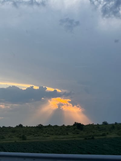 The image captures a rural landscape in Rupkite, Bulgaria, under a dynamic sky at either sunrise or sunset. The upper half of the frame is dominated by an overcast sky composed of varying shades of blue-grey and dark grey clouds. A prominent, elongated band of dark cumuliform clouds stretches horizontally across the middle of the sky. Above this central cloud formation, a thin, horizontal sliver of intense golden-yellow light is visible, suggesting direct sunlight. Below the main cloud band, on the right side of the frame, a distinct burst of vibrant golden-orange light emanates from behind the clouds, casting multiple, diverging crepuscular rays downwards towards the horizon.
The horizon line is occupied by a dark green, undulating landscape, heavily covered in dense vegetation, likely trees and shrubs, which appear silhouetted against the brighter light. In the immediate foreground, a flatter expanse of darker green, possibly agricultural fields with visible crop rows, is present. At the very bottom of the image, a horizontal, metallic grey structure, consistent with a guardrail or vehicle component, partially obstructs the view. No people or animals are visible in the scene. The overall atmosphere is one of natural grandeur, with the dramatic interplay of sunlight and clouds highlighting the rural environment.