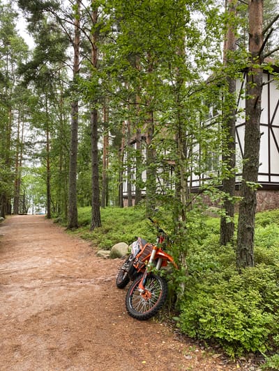 An orange dirt bike is parked on the side of a dirt path in a wooded area. The path leads into the distance, with trees on either side. A building with white siding and dark trim is visible behind the trees on the right side of the image. The ground is covered with dirt, pine needles, and low-lying green vegetation.