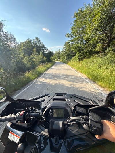 This is a first-person perspective image taken from an ATV on a paved road in Boltata, Bulgaria. The road stretches into the distance, flanked by lush green trees and tall grass. The sky is a clear blue with a single white cloud. The ATV's handlebars and dashboard are visible in the foreground, with the driver's hand on the right grip. The dashboard displays the number "37". The road appears to be in moderate condition with some cracks. The overall scene depicts an outdoor excursion on a sunny day.