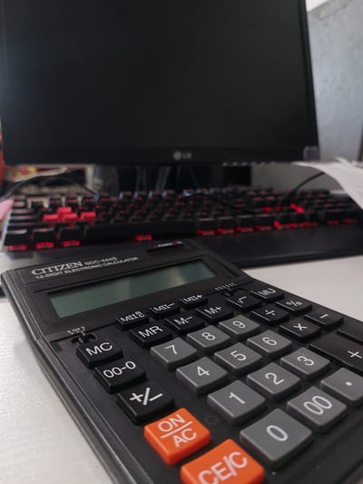This is a close-up, eye-level, static shot of a black Citizen SDC-444S electronic calculator positioned on a white surface. The calculator is the primary subject and takes up the majority of the foreground. Behind it, out of focus, is a black computer keyboard with red backlighting and a black computer monitor. The environment suggests an indoor workspace. The calculator has a display screen at the top, followed by a series of buttons including memory functions (M+, M-, MR, MC), square root, percentage, division, multiplication, subtraction, addition, and a numerical keypad from 0 to 9. There are also orange "ON/AC" and "CE/C" buttons at the bottom. The overall scene is well-lit and the focus is sharp on the calculator. There are no people or animals visible in the image. The location of Алфёрово, Russia is not discernible from the visual information presented in the image.