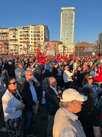 A large crowd of people, mostly adults, are gathered in an open area in İzmir, Türkiye, at the coordinates 38.436 latitude, 27.14 longitude. The crowd is standing on a grassy patch of land surrounded by buildings, including a tall building with "BOS PHALAS" written on it. The people are holding Turkish flags, with some people waving them. The crowd is facing a stage, and people are standing on the stage holding up banners. The people in the crowd are looking towards the stage, some are talking to each other, and some are waving their flags. The sky is blue and the sun is shining. The scene appears to be an outdoor political rally or demonstration.