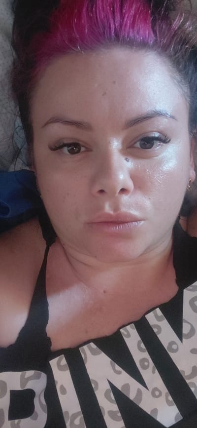 This is a close-up, head-and-shoulders selfie of a woman in what appears to be a bedroom setting. The woman has dark hair with bright pink highlights. She is wearing a black top with a leopard print pattern on the front that says "PINK". The woman has dramatic false eyelashes and appears to have some makeup on, including lip gloss. Her skin has a dewy or oily sheen to it. The background is soft and out of focus, with a hint of a grey textured surface behind her head and a blue object to her left. There is a small gold hoop earring visible in her right ear. The image was taken in Marynivka, Ukraine.