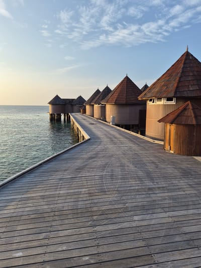 The image depicts a series of wooden overwater bungalows in Bodufolhudhoo, Maldives. The bungalows have peaked roofs and wooden siding. They are connected by a wooden boardwalk that extends over the water. The water is calm and reflects the sky. The sky is light blue with some clouds.