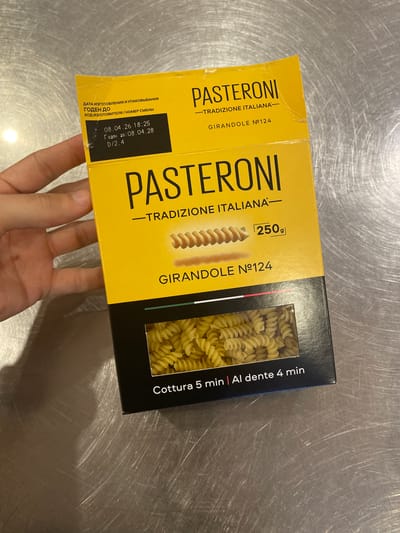 A person's hand is holding a yellow box of pasta. The box has the brand name "PASTERONI" printed in large black letters. Below the brand name, it says "TRADIZIONE ITALIANA" and "GIRANDOLE N°124". There is an image of fusilli pasta on the box, and a small rectangle indicating "250g". A window in the box reveals the actual pasta inside. At the bottom of the box, text reads "Cottura 5 min | Al dente 4 min". A label on the top left of the box has Cyrillic text that translates to "Date of manufacture and packaging", "BEST BY", and "Manufacturer code/shift number". Below this are the dates "08.04.26 18:25" and "BEST BY 08.04.28". The box is placed on a metallic, brushed surface. The lighting suggests an indoor environment, possibly a kitchen or pantry, and the surface indicates a countertop. There is no discernible information in the image to indicate a specific location in the city of Oryol, Russia.