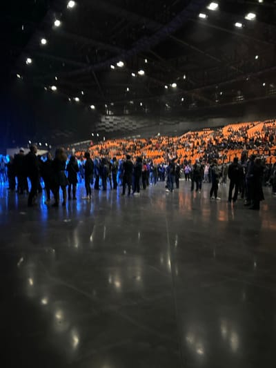 An indoor venue features rows of orange seating filled with people. A line of individuals stands on a dark, reflective surface, their forms blurred and illuminated by a blue light. Above, a high ceiling is visible, structured with metal beams and dotted with evenly spaced lights. The lighting casts elongated reflections on the shiny floor. The image has a strong vertical orientation.