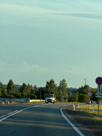The image shows a two-lane road in a rural setting, likely near Togashevo, Russia, given the provided context. The road curves to the right, with a white lane divider. On the right side of the road, there are several traffic signs. One sign, at the top, is a circular red sign with a blue cross inside, indicating no stopping. Below it are two circular signs, one with two cars side-by-side indicating a passing zone, and another with "70" suggesting a speed limit. Further down, there is a sign depicting a tow truck, likely indicating a towing zone or service. The road is bordered by a grassy shoulder and a low white fence. Beyond the fence, there are trees and some houses visible in the distance. A white van is driving away from the camera on the right lane, and two cars are parked on the right side of the road. The sky is a pale blue with scattered clouds, and the sunlight suggests it is either early morning or late afternoon. The overall scene depicts a typical roadside environment in a suburban or rural area.