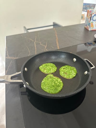 Three bright green pancakes are being cooked in a black non-stick frying pan, which is placed on a black induction stovetop. The pancakes have a slightly uneven, bubbly surface, indicating they are in the process of cooking. The frying pan has a silver handle with the word "NINJA" visible on it. The stovetop is set on a dark, marble-patterned countertop. In the background, a tissue box with a colorful floral pattern is visible on the countertop, and a white wall can be seen. The scene appears to be a kitchen environment. There are no people visible in the image. This image was taken in Leeds, United Kingdom.