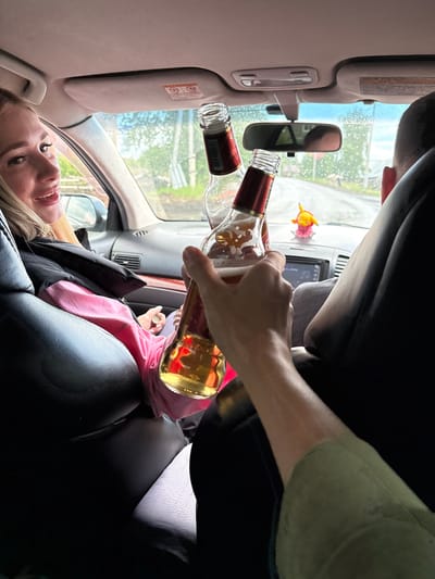 Two people are inside a car, likely in the city of Zheleznodorozhny, Russia, on a rainy day. The woman in the passenger seat is looking towards the camera with a smile, her hair blonde and pulled back. She is wearing a black vest over a pink hoodie and a light-colored garment underneath. In the foreground, a person's hand is holding up two bottles of beer, one of which is partially filled. The bottles have red and gold labels. The interior of the car appears to be a sedan, with leather seats. A rearview mirror is visible, reflecting the rainy road ahead. A small yellow Pikachu figurine is perched on the dashboard. The overall scene suggests a casual outing, possibly a drive with friends.