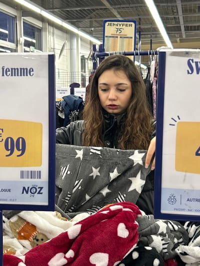 A young woman with long, curly brown hair is shopping in a store. She's wearing a black jacket and appears to be looking at a pile of cozy blankets with star and heart patterns. Price signs with "femme €9.99" and "JENYFER €75" are visible, indicating the location is a clothing or department store. The background shows clothing racks and store lighting.