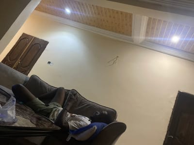 This image, taken from a low-angle perspective and rotated counter-clockwise, depicts an indoor domestic scene. The primary subject is a person, appearing to be an adult male, lying on their back on a dark upholstered sofa or bed. They are dressed in a white short-sleeved t-shirt and green or camouflage shorts, with their head resting on a blue cushion. Their arms are bent across their body, and their legs are also bent, suggesting they are resting or sleeping. A clear plastic bag is partially visible near the person's feet on the furniture.

The shot looks upwards towards a light-colored, possibly cream or off-white, ceiling. Several notable features are visible on the ceiling: an exposed bundle of electrical wires hangs loosely from a central point, and a wall outlet or light switch is mounted on the ceiling further to the right. Two rectangular wooden panels, possibly access hatches or decorative elements, are integrated into the ceiling; one is in the upper left corner of the frame, and another is in the mid-right.

Below the ceiling, a decorative trim runs along the top of the wall, which is adorned with wood-grain patterned paneling or wallpaper. Bright reflections, likely from windows or artificial light sources, are visible on these paneled surfaces, indicating illumination in the room. The overall setting appears to be a modest interior, possibly a living room or bedroom, situated in Lakka, Sierra Leone. No visible text is present in the image.