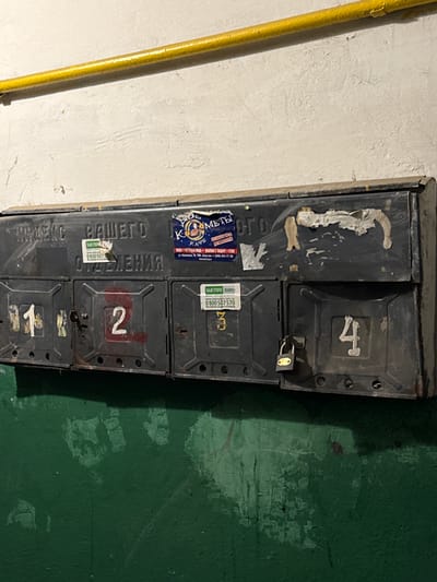 A weathered, dark metal mailbox unit is mounted on a textured, off-white wall with a bright yellow pipe running horizontally across the top. The mailbox has multiple compartments, four of which are clearly visible. These compartments have numbers painted on them: "1" on the first, "2" on the second, "3" on the third, and "4" on the fourth. The mailbox is adorned with various stickers and peeling paper, some with text and graphics, including what appears to be a logo with a bird and text in Cyrillic. The third compartment from the left has a small, silver padlock attached. The bottom of the image shows a rough, dark green painted wall with areas of chipping and wear. The scene appears to be an interior hallway or common area within a building.